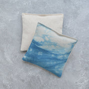 Two square fabric lavender sachets on a textured gray surface. The front bean bag has a blue and white watercolor sky print, while the back bean bag is a natural cream linen color.