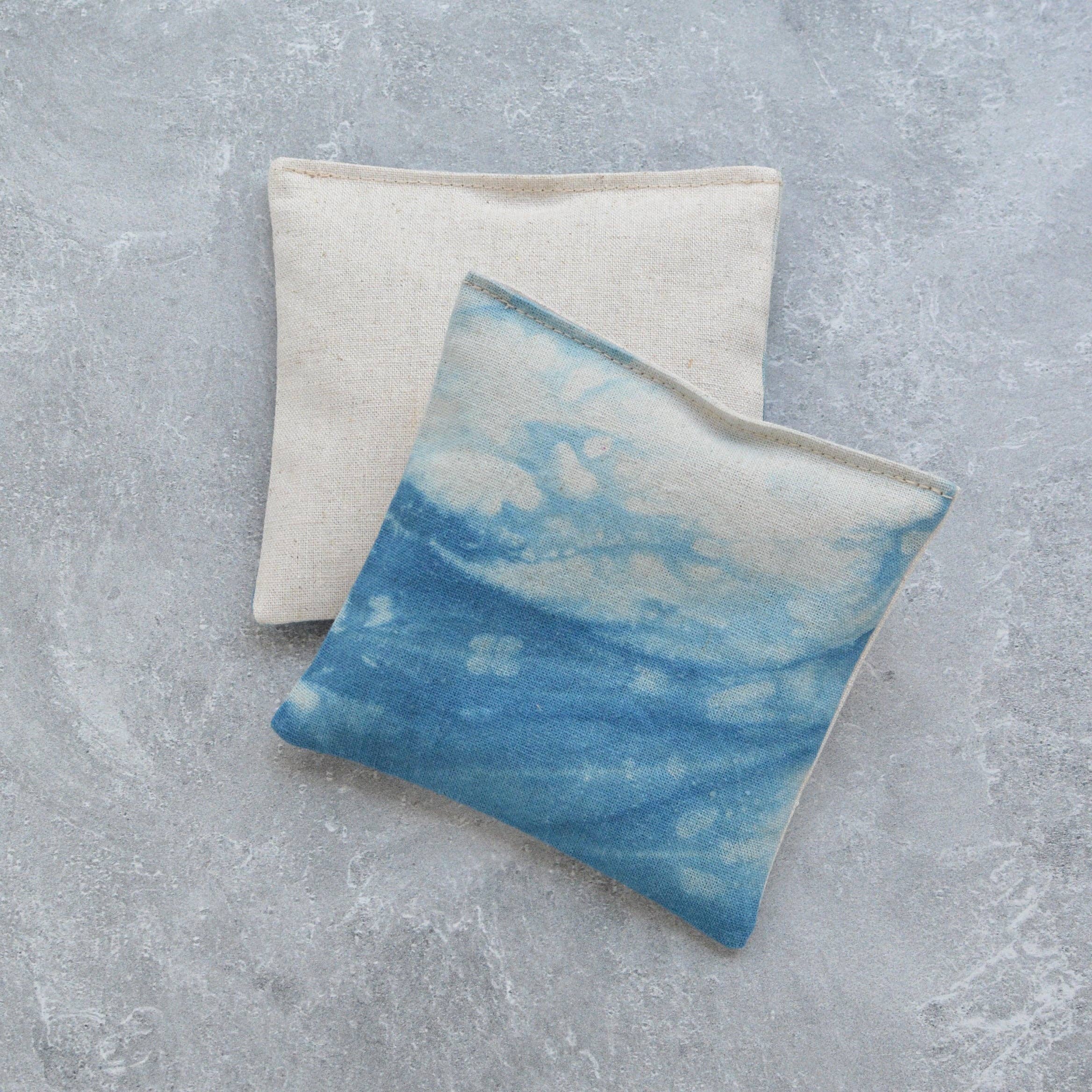 Two square fabric lavender sachets on a textured gray surface. The front bean bag has a blue and white watercolor sky print, while the back bean bag is a natural cream linen color.