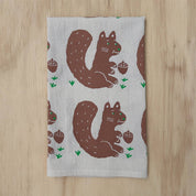 A rectangular tea towel with a repeating pattern of brown squirrels holding acorns on a light gray background, laid flat on a light wood surface.