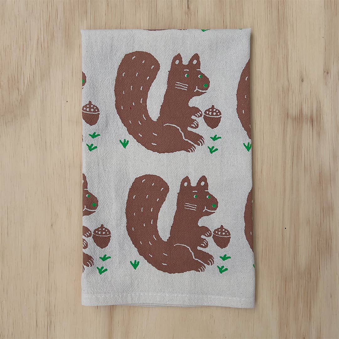 A rectangular tea towel with a repeating pattern of brown squirrels holding acorns on a light gray background, laid flat on a light wood surface.