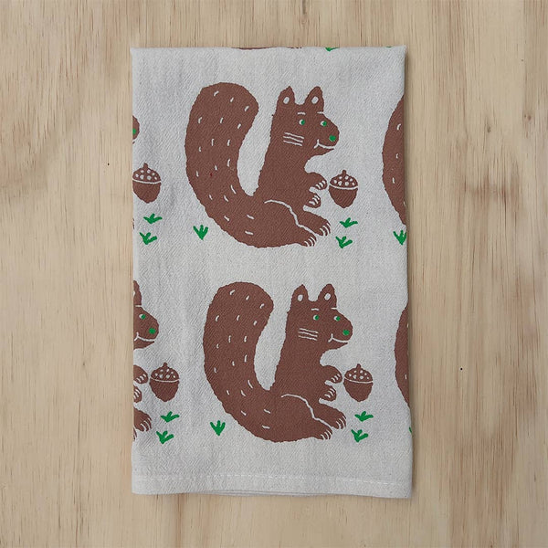 A rectangular tea towel with a repeating pattern of brown squirrels holding acorns on a light gray background, laid flat on a light wood surface.