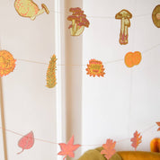 An autumn-themed paper garland hanging in a room, featuring colorful paper leaves, flowers, acorns, and mushrooms strung along thin threads.
