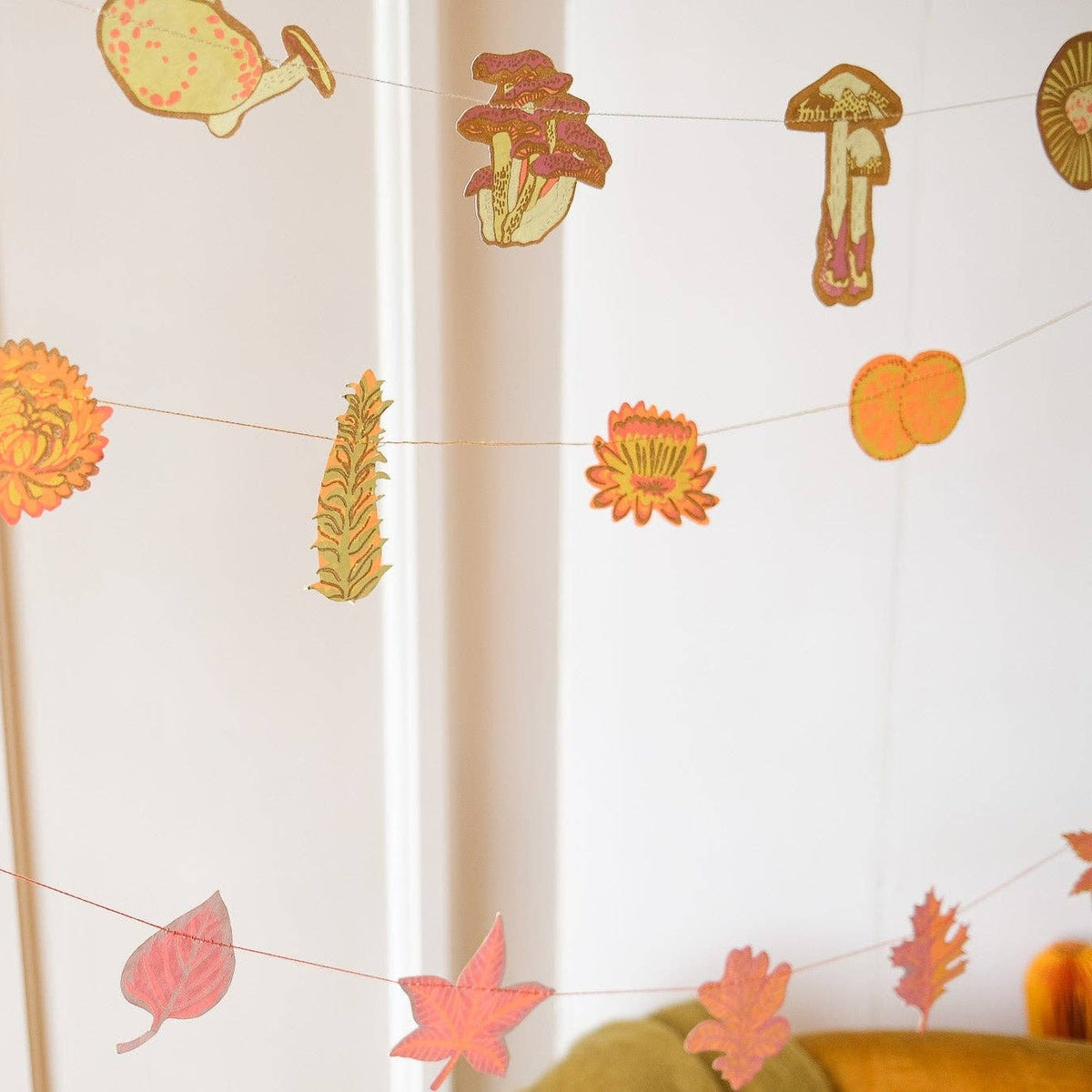 An autumn-themed paper garland hanging in a room, featuring colorful paper leaves, flowers, acorns, and mushrooms strung along thin threads.