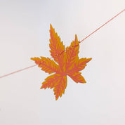 An orange and yellow autumn maple leaf hanging from a thin string against a pale gray background.