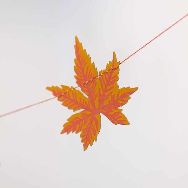 An orange and yellow autumn maple leaf hanging from a thin string against a pale gray background.