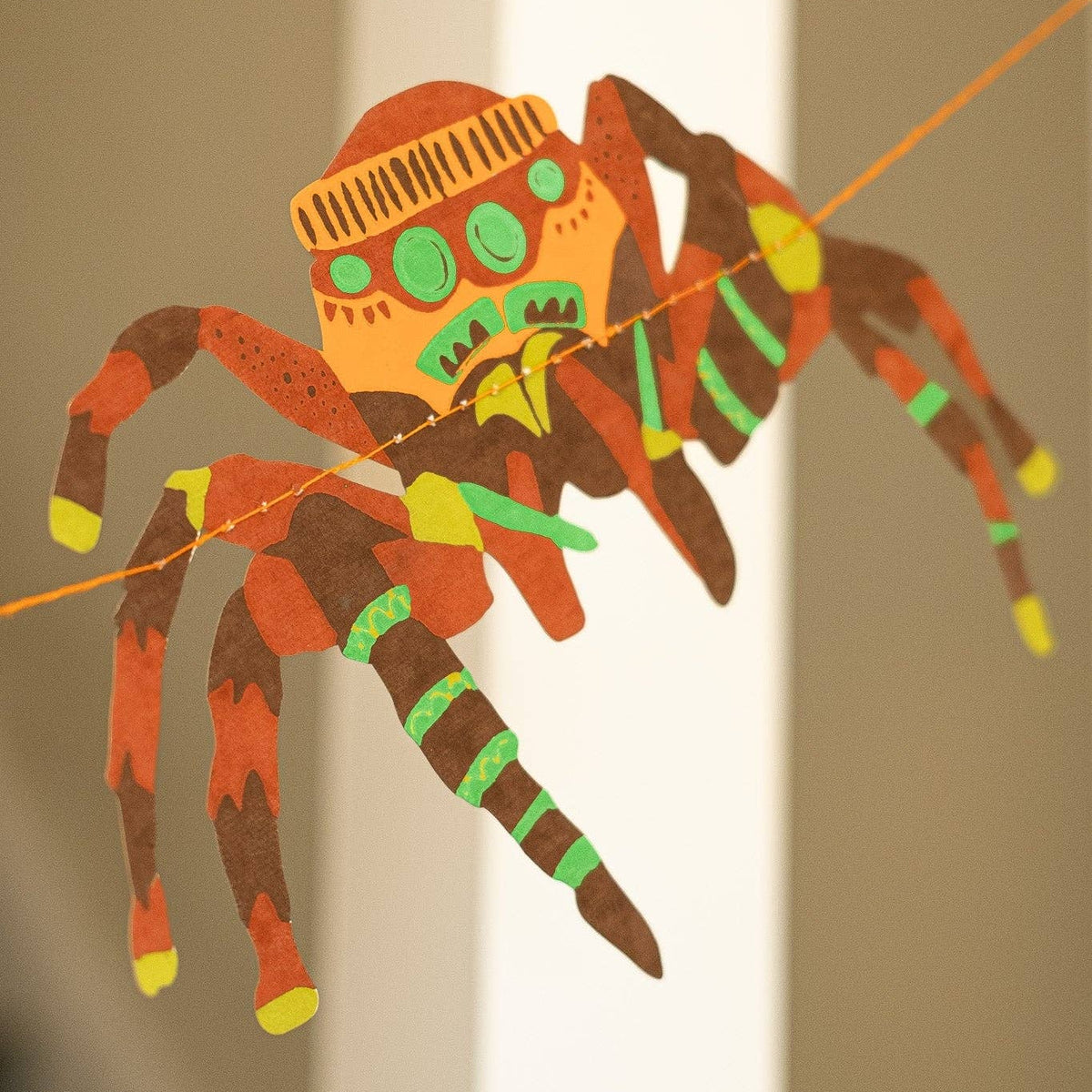 A colorful handmade spider decoration made from patterned paper, strung along a thin thread against a neutral indoor background.