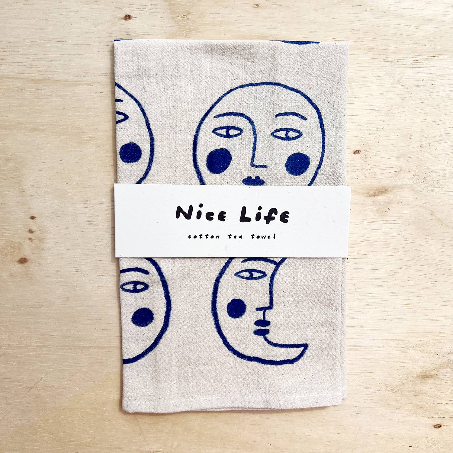 A cotton tea towel with face designs on a wooden surface, branded 'Nice Life'.
