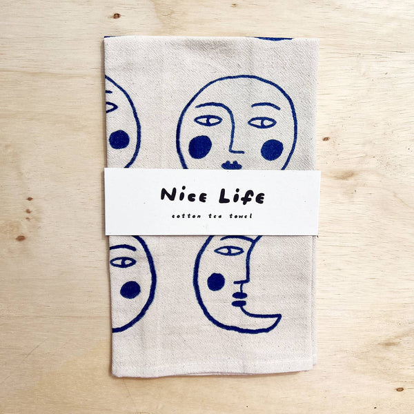 A cotton tea towel with face designs on a wooden surface, branded 'Nice Life'.