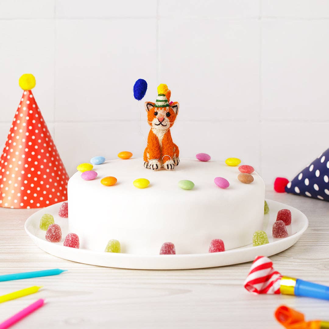 White cake with a orange cat topper and colorful candies on a white plate, surrounded by party hats and candles.
