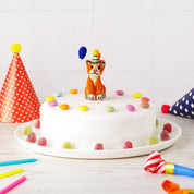 White cake with a orange cat topper and colorful candies on a white plate, surrounded by party hats and candles.