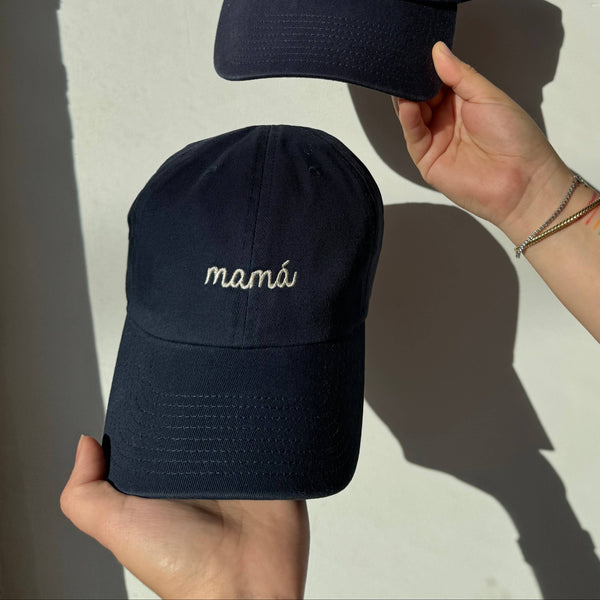 Two navy blue caps with "mamá" text held by a person against a light background