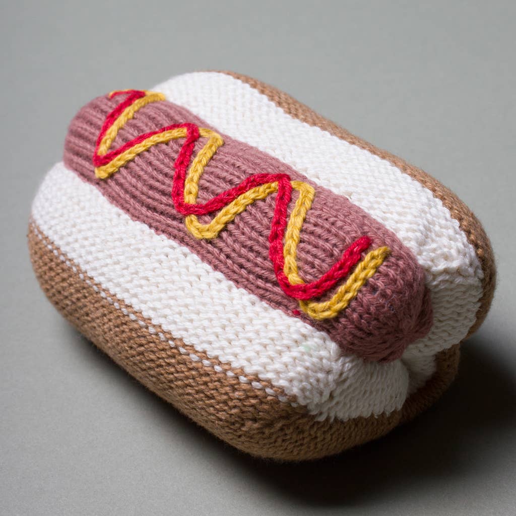 A knitted hot dog toy or plush accessory on a smooth gray background. The hot dog features a white bun with a tan crust, a pinkish-brown sausage, and yellow and red wavy accents representing mustard and ketchup.