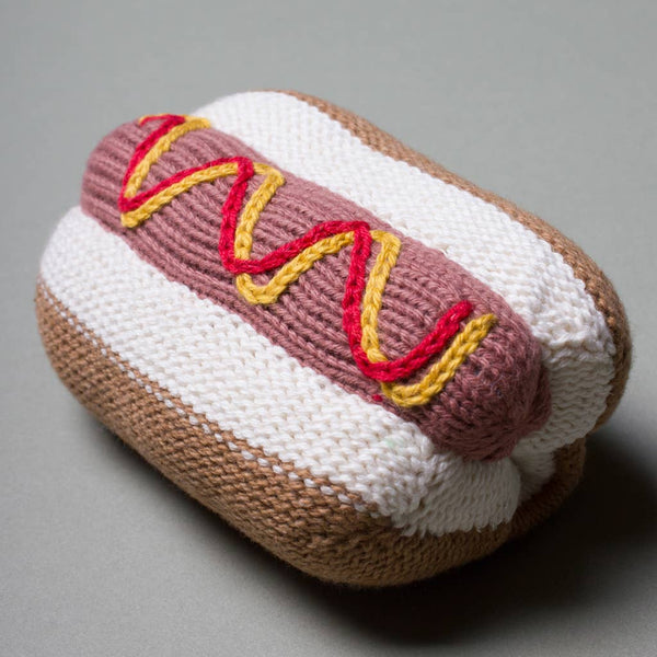A knitted hot dog toy or plush accessory on a smooth gray background. The hot dog features a white bun with a tan crust, a pinkish-brown sausage, and yellow and red wavy accents representing mustard and ketchup.