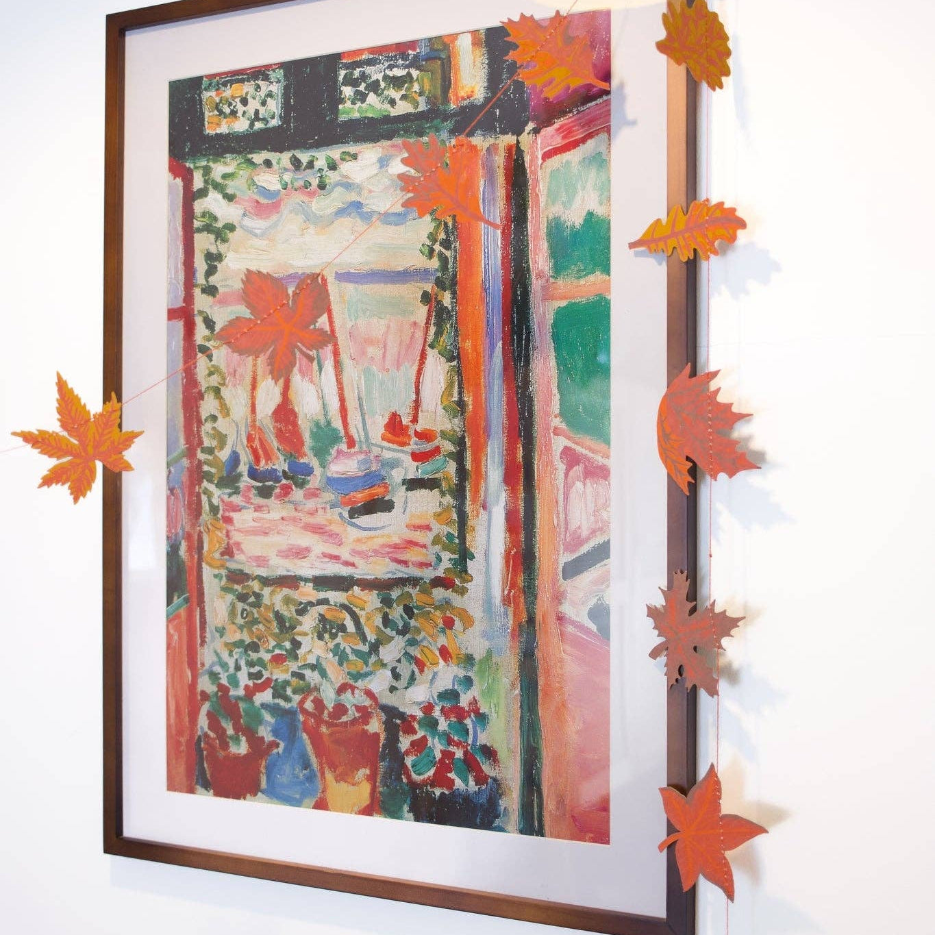 A framed impressionist-style painting hangs on a white wall, decorated with orange paper maple leaves strung along the top and left edges of the frame.
