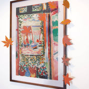 A framed impressionist-style painting hangs on a white wall, decorated with orange paper maple leaves strung along the top and left edges of the frame.