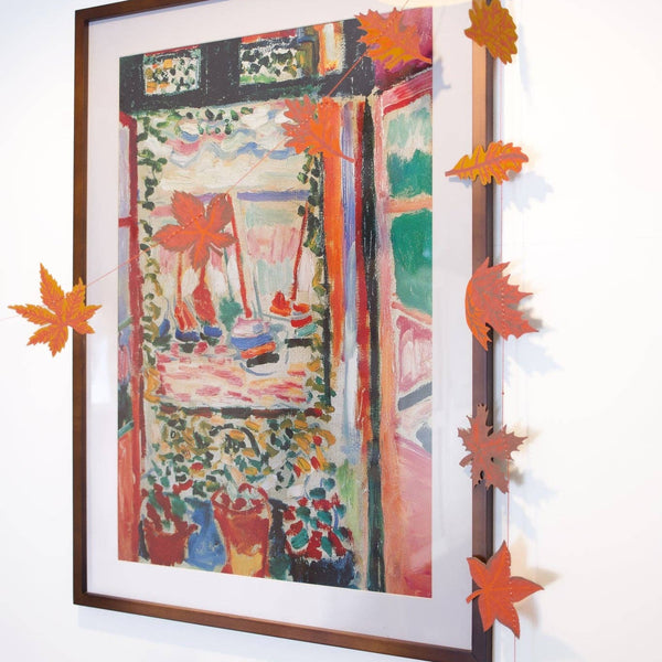 A framed impressionist-style painting hangs on a white wall, decorated with orange paper maple leaves strung along the top and left edges of the frame.