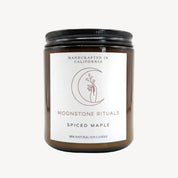A brown glass jar candle with a black metal lid and a white label from Moonstone Rituals, labeled 'Spiced Maple' and '100% Natural Soy Candle'.