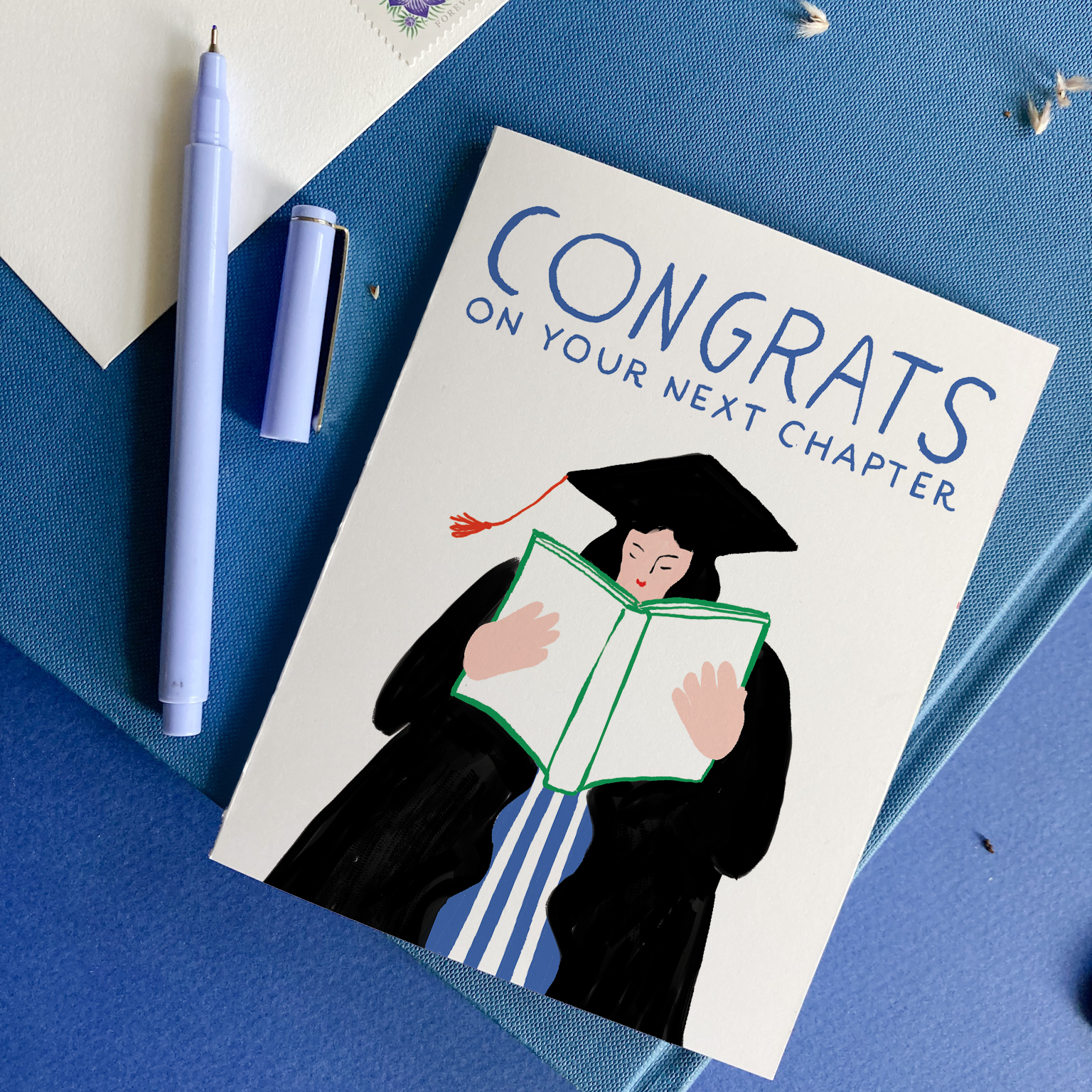 Graduation-themed card with a person in a cap and gown reading a book, on a blue surface with a pen and small card.