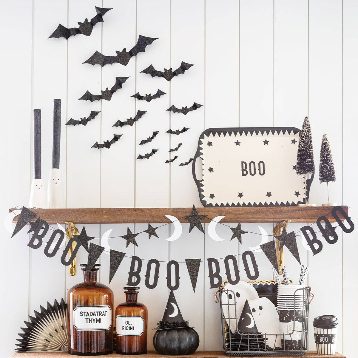 Halloween-themed shelf decor arrangement on two wooden floating shelves against a white shiplap wall, featuring black bat cutouts, a 'BOO' banner, star and moon garlands, amber bottles, a small black pumpkin, a tray sign, and spooky accessories.