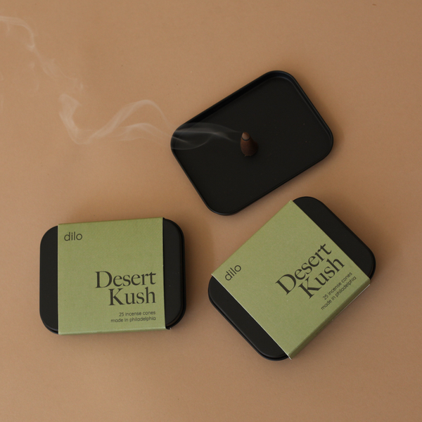 Two black containers labeled 'Desert Kush' with a small incense stick on a beige background.