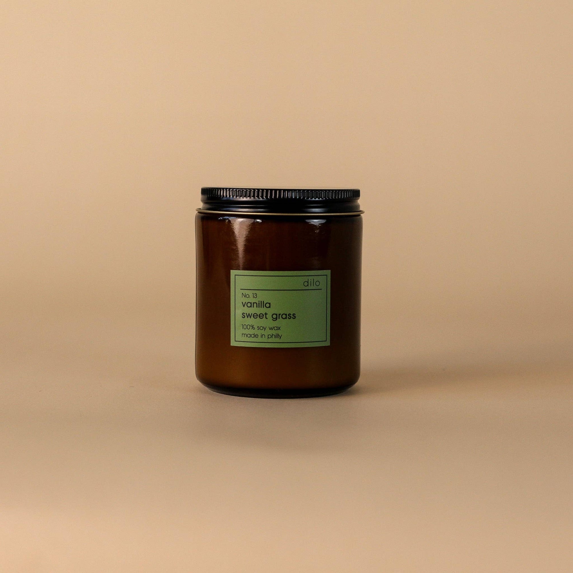 A brown glass candle jar with a black lid, featuring a label that reads 'Vanilla Sweet Grass'. The jar is set against a beige background.