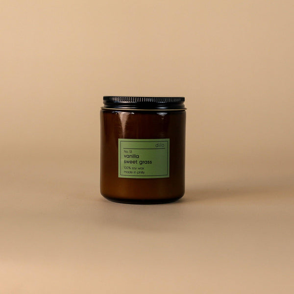 A brown glass candle jar with a black lid, featuring a label that reads 'Vanilla Sweet Grass'. The jar is set against a beige background.