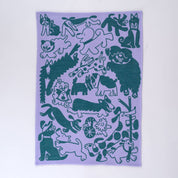 A rectangular woven tea towel, featuring a playful pattern of various cartoon animals in dark teal on a light lavender background, shown flat against a white backdrop.