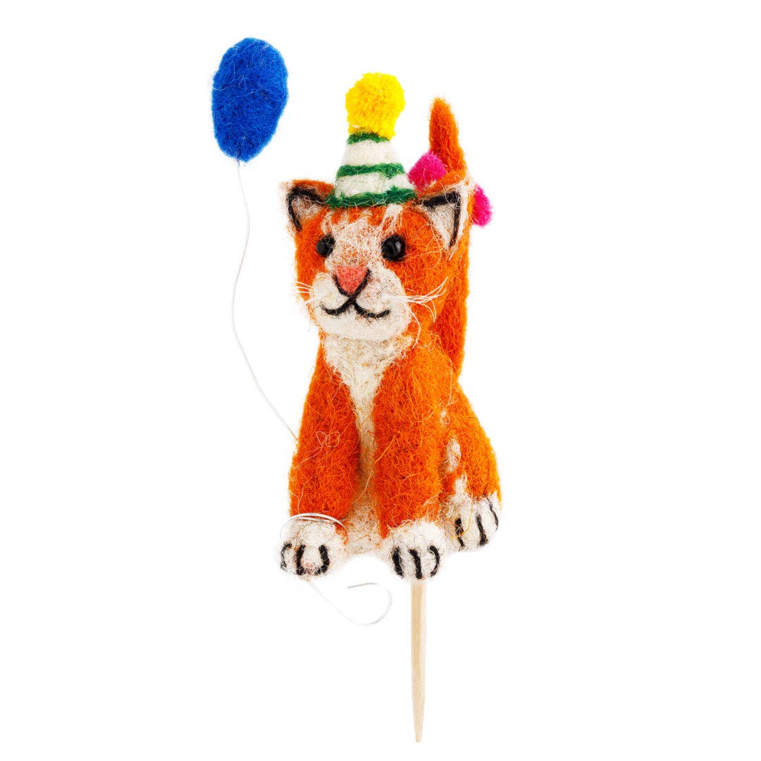Felted cat cake topper with colorful hat and pom-poms on a white background