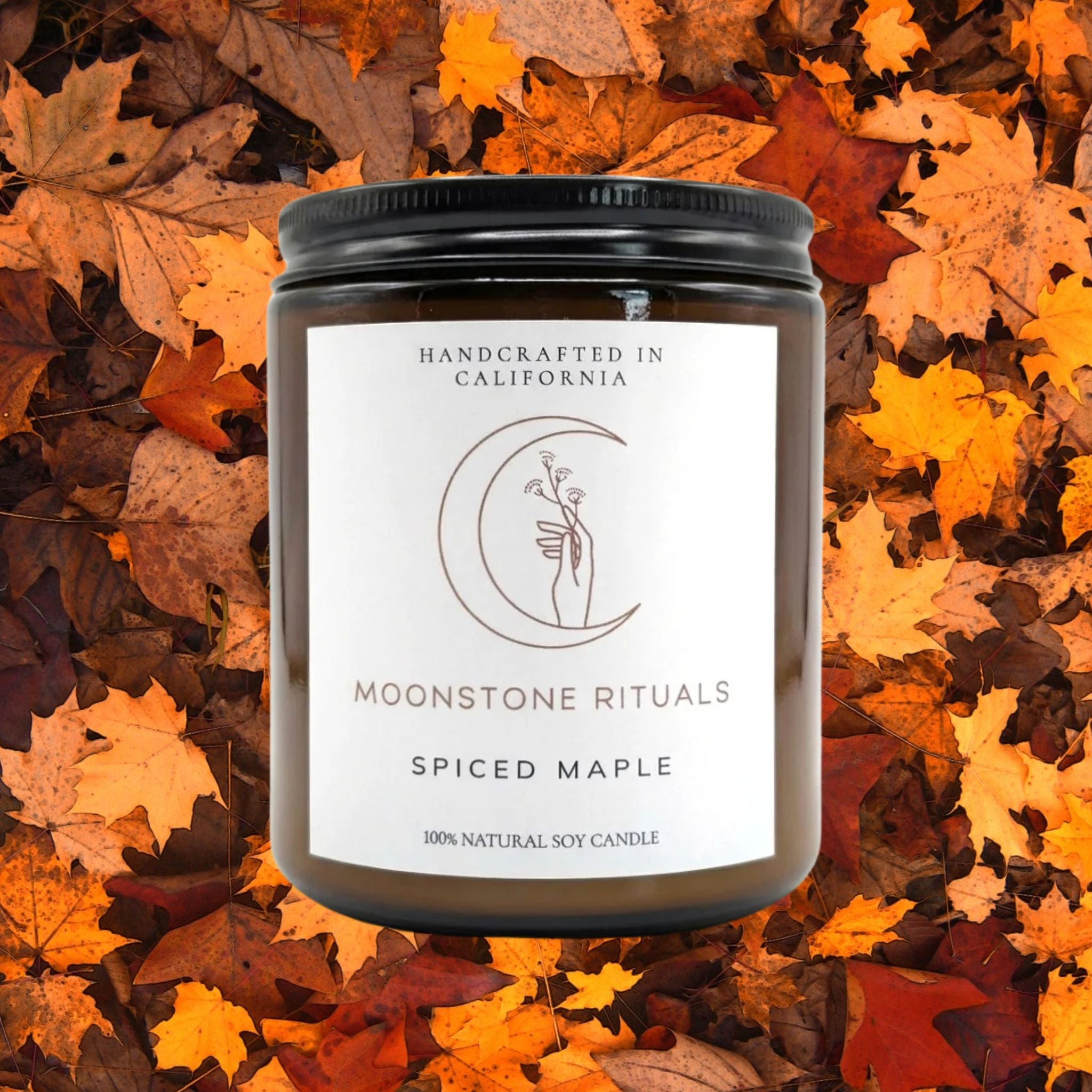 A brown glass jar candle with a black metal lid and a white label from Moonstone Rituals, labeled 'Spiced Maple' and '100% Natural Soy Candle'.