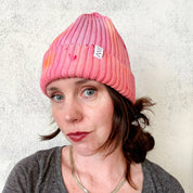 Person wearing a pink beanie with a brand logo against a textured white wall