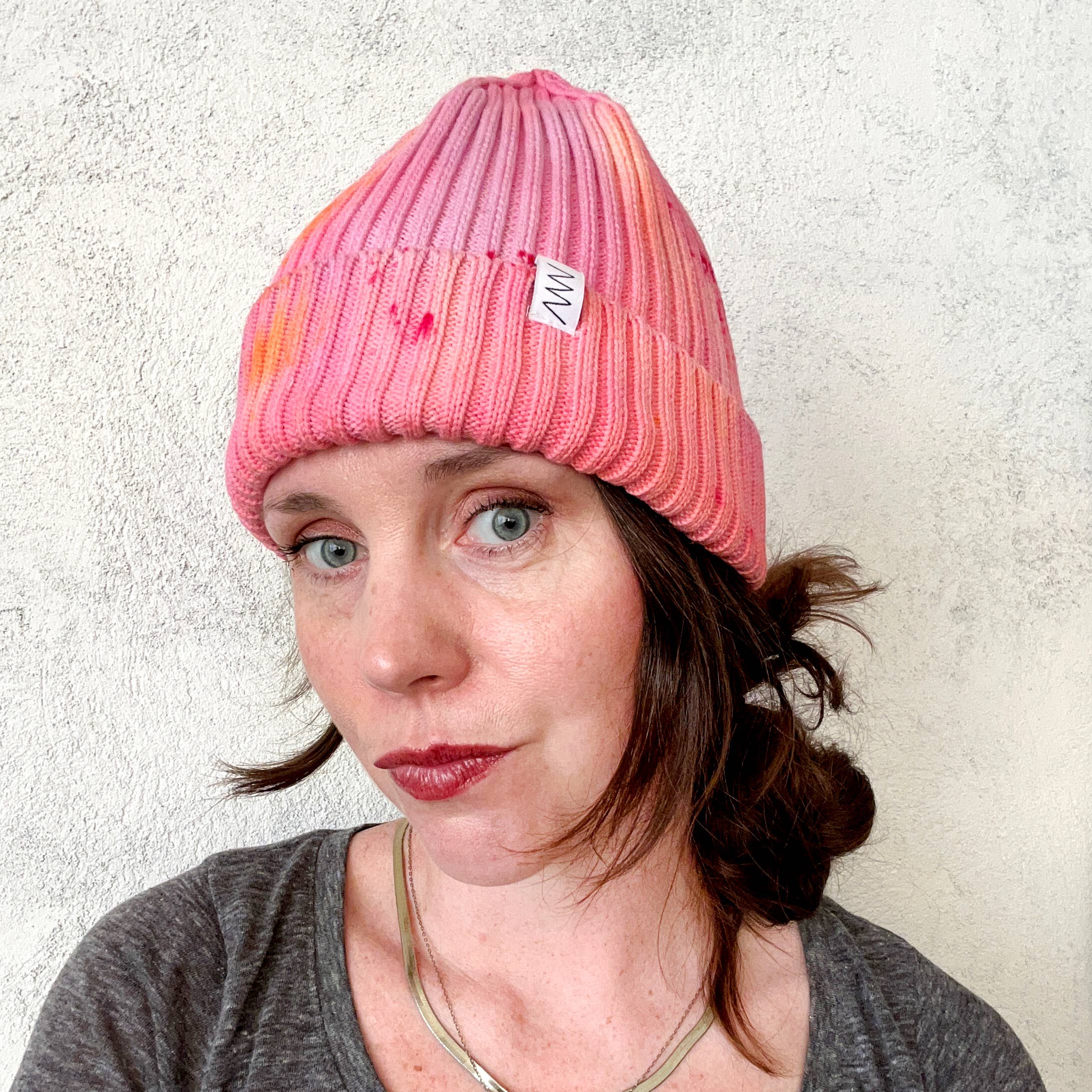 Person wearing a pink beanie with a brand logo against a textured white wall