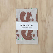 A folded cotton tea towel with a brown squirrel and acorn pattern on a light beige background, wrapped with a white paper band that reads 'Nices Life cotton tea towel', placed on a light wood surface.