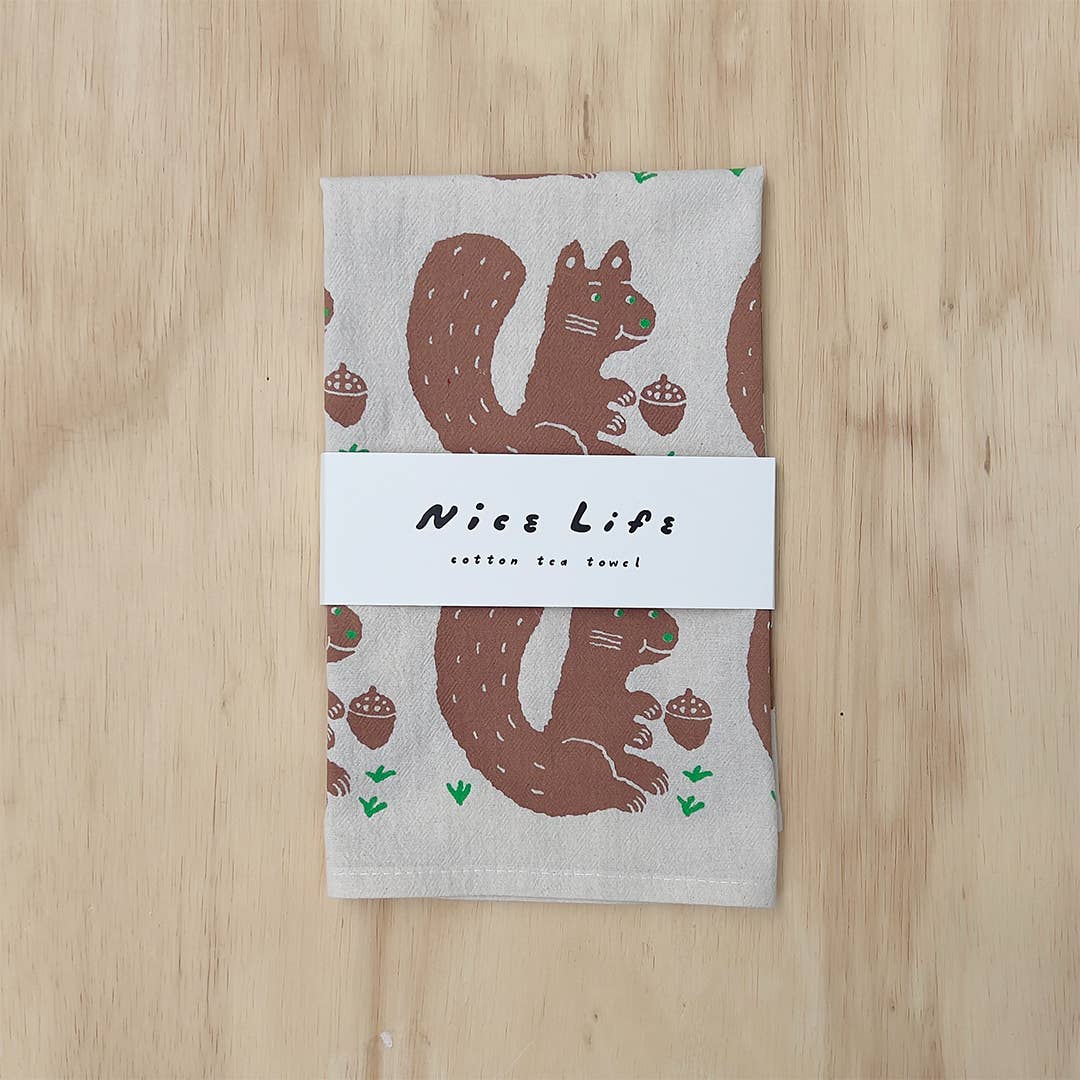 A folded cotton tea towel with a brown squirrel and acorn pattern on a light beige background, wrapped with a white paper band that reads 'Nices Life cotton tea towel', placed on a light wood surface.