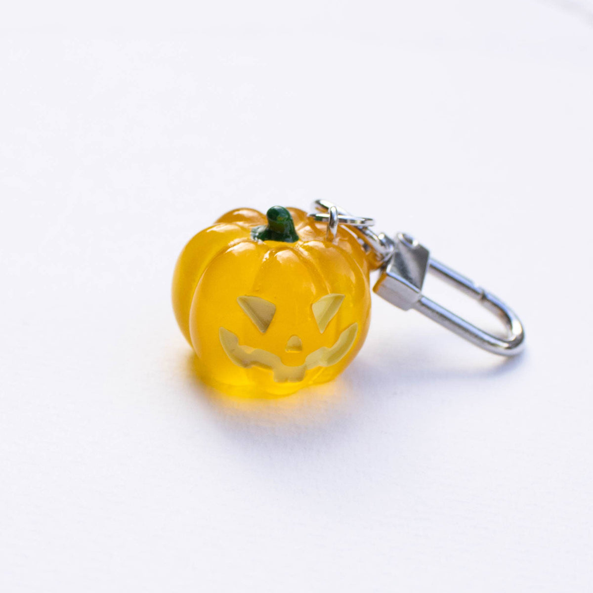A small translucent orange jack-o'-lantern charm with a carved face and a silver metal lobster clasp, resting on a plain white surface.
