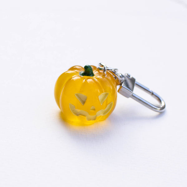 A small translucent orange jack-o'-lantern charm with a carved face and a silver metal lobster clasp, resting on a plain white surface.