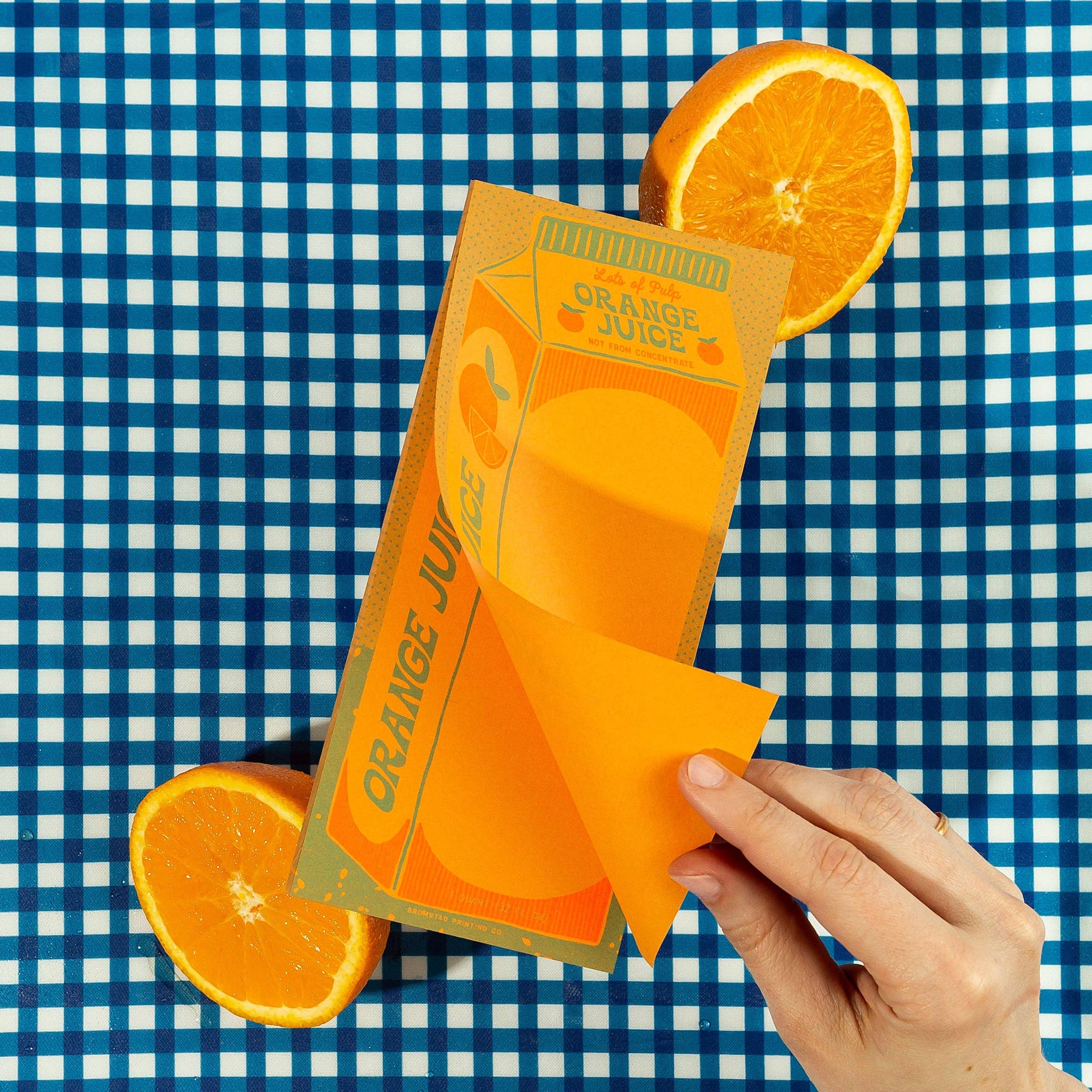 A note pad designed to look like a carton of orange juice with a sliced orange on a blue and white checkered background. A hand is holding the top sheet of the pad.