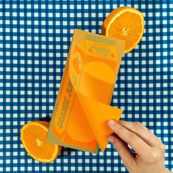 A note pad designed to look like a carton of orange juice with a sliced orange on a blue and white checkered background. A hand is holding the top sheet of the pad.