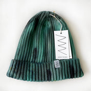 A ribbed knit beanie hat in a variegated green and teal colorway with dark charcoal accents, featuring a white paper hangtag and a small woven brand label on the cuff. The brand 'Merle Works' is visible on the tag.