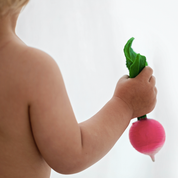 A close-up lifestyle shot of a baby's arm and shoulder holding a bright pink and green radish-shaped toy against a soft white background.