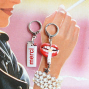 Two novelty earrings displayed on a hand against a pink and yellow illustrated background. One earring has a silver ring and a white lighter charm with the red word 'merci'.