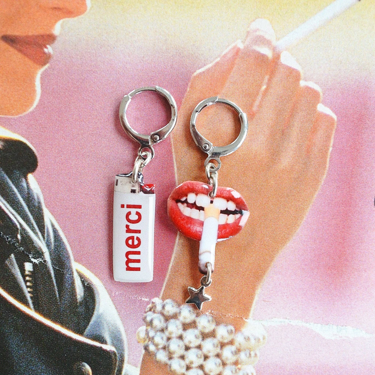 Two novelty earrings displayed on a hand against a pink and yellow illustrated background. One earring has a silver ring and a white lighter charm with the red word 'merci'.