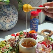 A hand holds a wooden honey dipper dripping honey over a small white bowl on a charcuterie board, with a jar of Zab's Hot Honey in the background.
