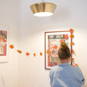 A woman in a light blue shirt is seen from behind, hanging orange paper leaves along a string between two framed art prints on a white wall. A gold pendant light hangs from the ceiling, and a wooden shelf with a green plant is visible on the left.