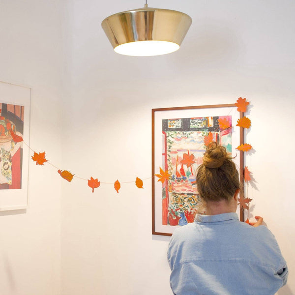 A woman in a light blue shirt is seen from behind, hanging orange paper leaves along a string between two framed art prints on a white wall. A gold pendant light hangs from the ceiling, and a wooden shelf with a green plant is visible on the left.
