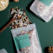 Three clear stand-up pouches of Ritual No. 8 Winter Sanctuary bath tea are arranged on a dark green surface with decorative gold ornaments, pinecones, and spilled loose tea and sugar.