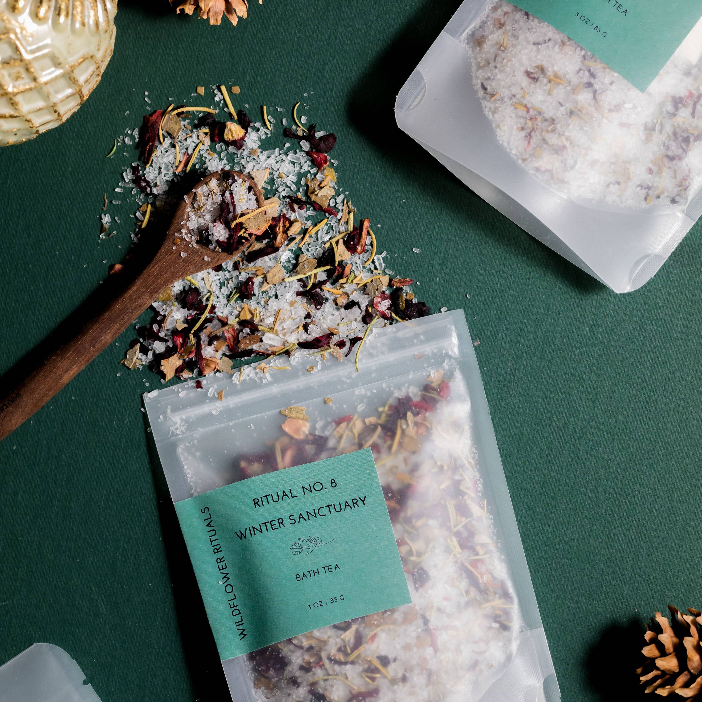 Three clear stand-up pouches of Ritual No. 8 Winter Sanctuary bath tea are arranged on a dark green surface with decorative gold ornaments, pinecones, and spilled loose tea and sugar.