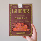 A hand holds a box of East End Press Garland printed paper against a plain light gray background. The box is dark reddish-brown with gold decorative border text and a clear window showing orange-red paper leaves inside.