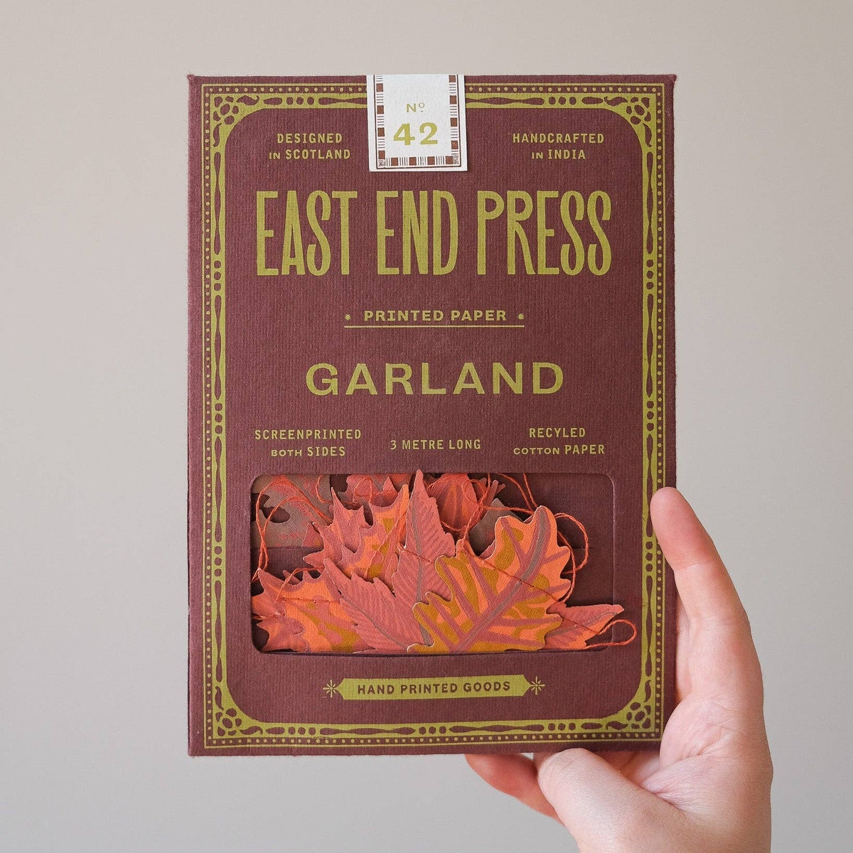 A hand holds a box of East End Press Garland printed paper against a plain light gray background. The box is dark reddish-brown with gold decorative border text and a clear window showing orange-red paper leaves inside.