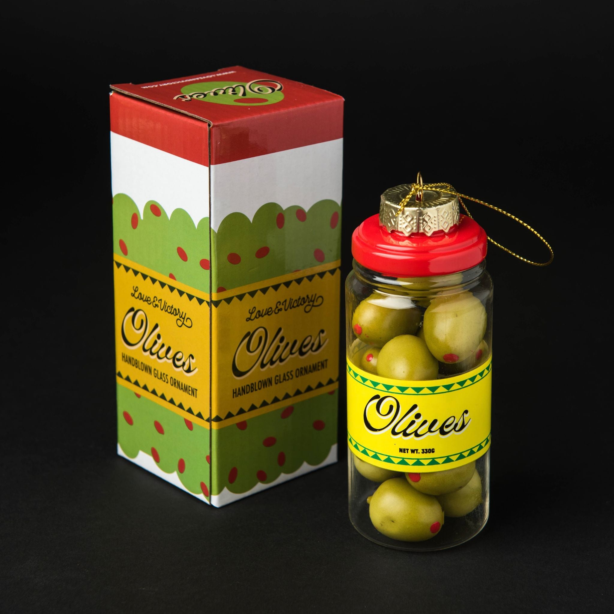 A jarred olive ornament displayed next to its retail box on a black background. The clear glass jar contains green olives and has a red lid with a gold hanging ornament loop and bow.
