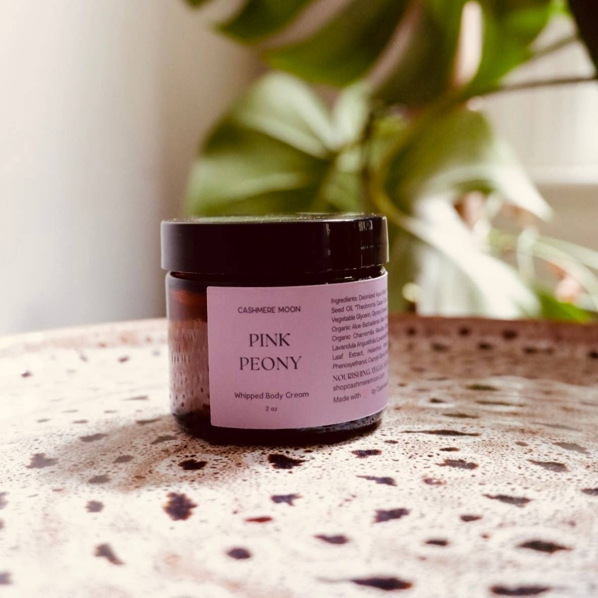 Jar of Pink Peony whipped body cream on a textured surface with a plant in the background